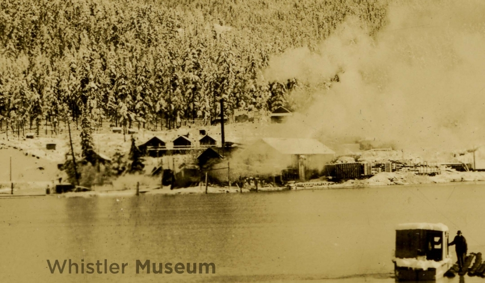03Support image_Whistler Museum story_Parkhurst Mill in 1935_Photo Rowland Collection | Trans Canada Trail Old sepia photo of a smoky lakeside town with trees on a hill and a docked vehicle; Whistler Museum watermark