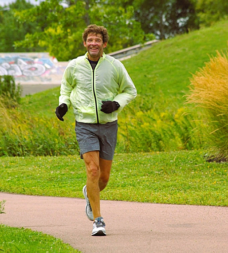 05Support image_John Dallaire_credit Cathy Donaldson | Trans Canada Trail Man jogging on a park path wearing a light green jacket, black gloves, and gray shorts, with trees and grass in the background.