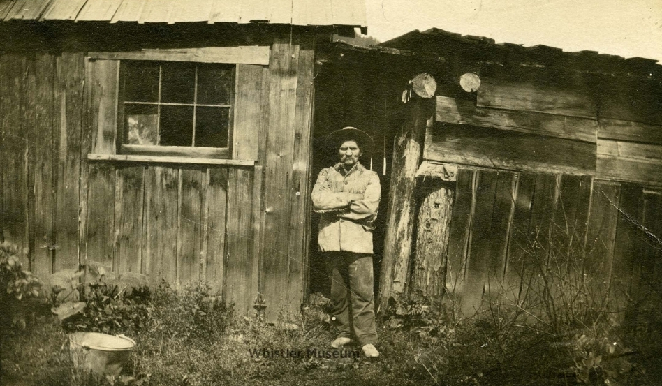 05Support image_Whistler story_John Millar outside his cabin_credit Whistler Museum-¨hillip Collection | Trans Canada Trail Older man with arms crossed stands in front of a weathered wooden shed, a small window to his left and overgrown ground around him.