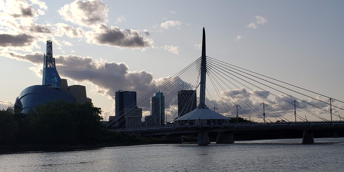 MB_City-of-Winnipeg_Esplanade_Web_StoryFeatured_Image_1200x600 | Trans Canada Trail Cable-stayed bridge with a tall central pylon spanning a calm river, set against a city skyline with a distinctive curved blue building on the left at dusk/light clouds above.