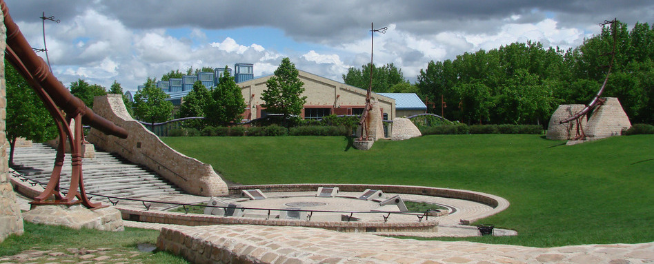 Outdoor sculpture garden with tall rust-colored metal sculptures around a circular stone plaza on a green lawn under cloudy skies.