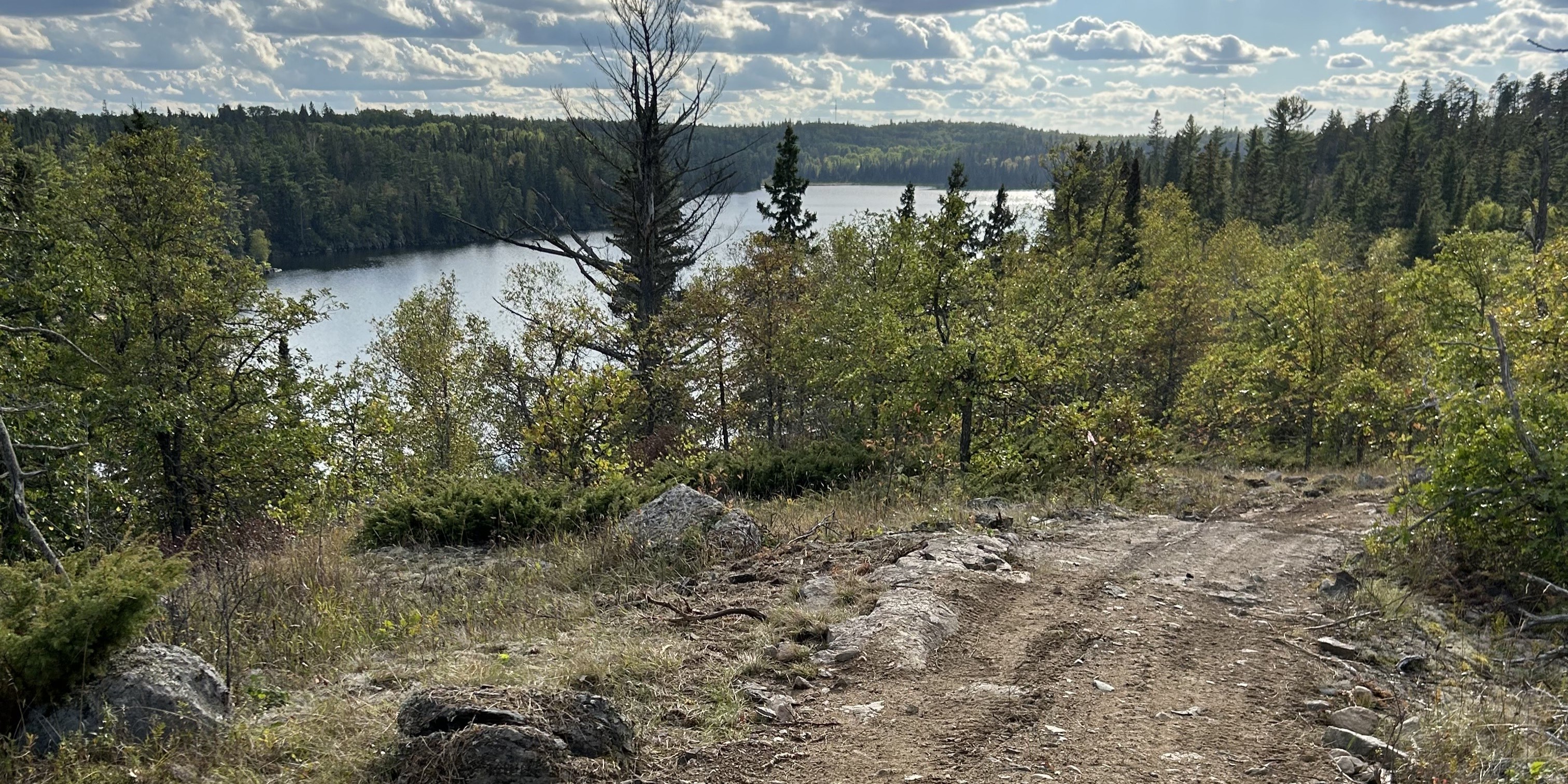 Dirt trail winding through rocks and shrubs toward a calm lake surrounded by dense evergreen forest under a partly cloudy sky?