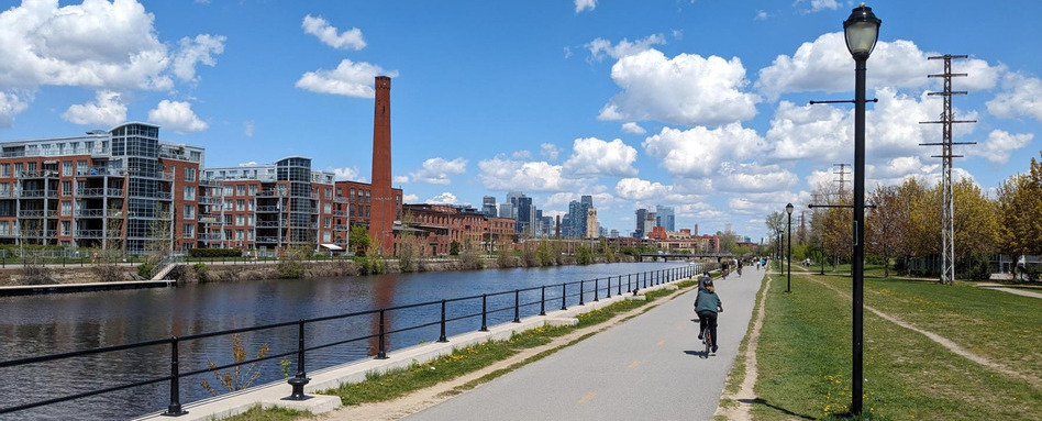 Riverside bike path with a cyclist beside the water, city buildings on the left and a distant skyline under a blue sky.