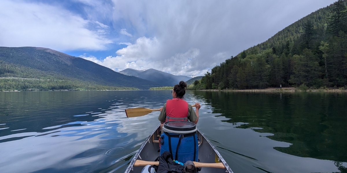 00Feature image_Ali Becker web story_West Arm Paddle Route | Trans Canada Trail Person wearing a pink life vest paddles a canoe across a calm mountain lake, with forested hills and mountains under a partly cloudy sky (back view).