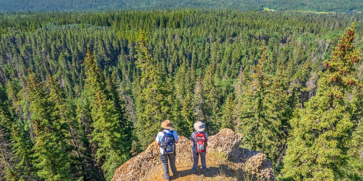 00Feature image_SK_Cypress HIlls Interprovincial Park_Credit Robin and Arlene Karpan | Trans Canada Trail Two people facing away from the camera, standing on a rocky overlook. Below them is a dense forest.