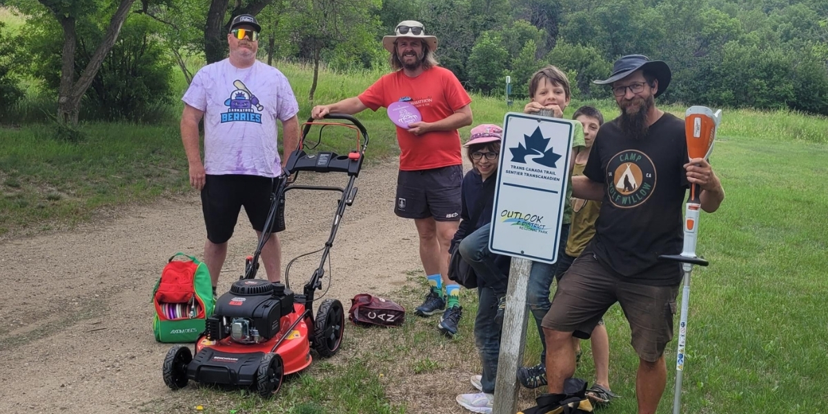 00Feature image_Trail Care 2025 lookback_credit Town of Outlook | Trans Canada Trail Five people on a dirt trail with a red lawn mower and hiking gear, standing beside a trail sign in a green field.