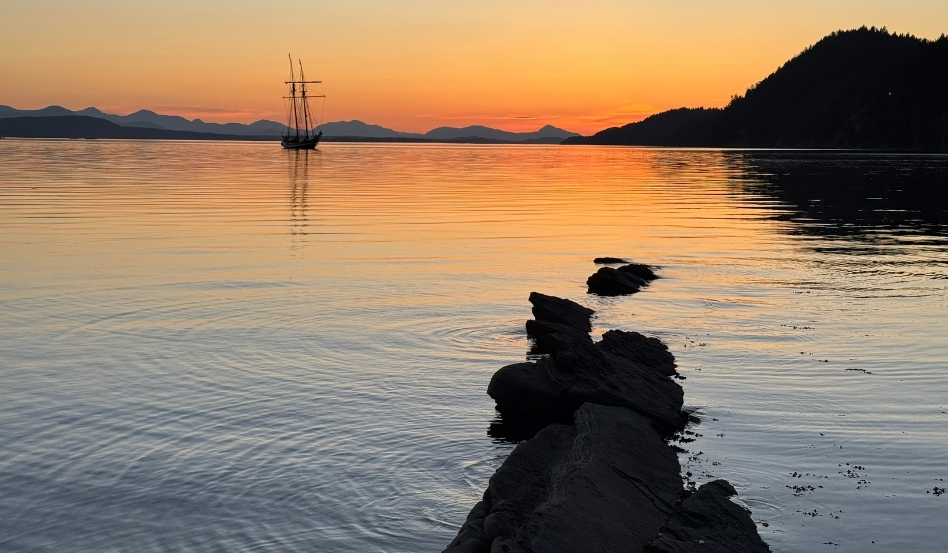Sunset over calm water with a single-masted sailboat near the horizon and a rocky jetty stretching into the foreground.