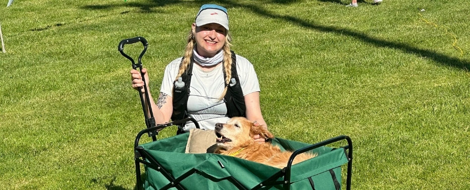 A woman smiles at the camera. She is crouching next to a wagon where an elderly dog is lying down.