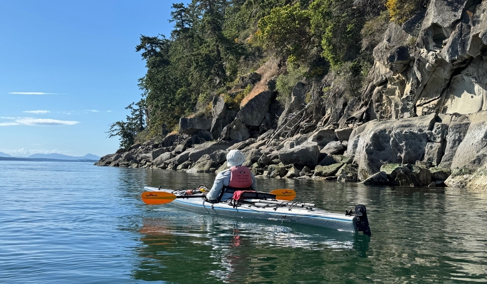Person kayaking in a calm bay beside a rocky cliff with trees, wearing a pink life jacket and white hat/ocean scenery behind.