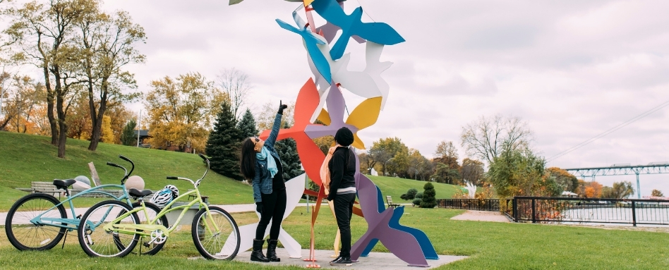 02Support image_ON_TWEPI article_Windsor sculpture garden_credit Tourism Windsor Essex Pelee Island | Trans Canada Trail people standing infront of a statue waterfront with bikes and bridge behind