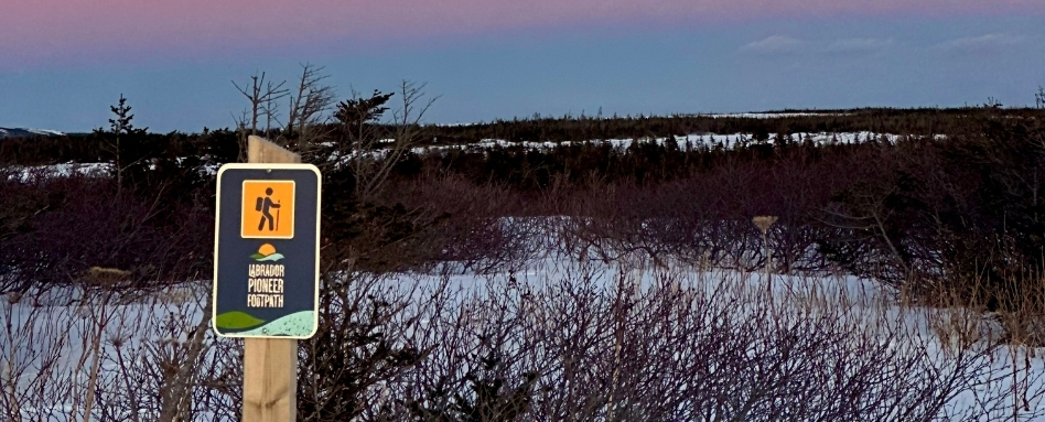 Trail sign on a wooden post in a snow-covered landscape at dusk; reads Labrador Pioneer Footpath.
