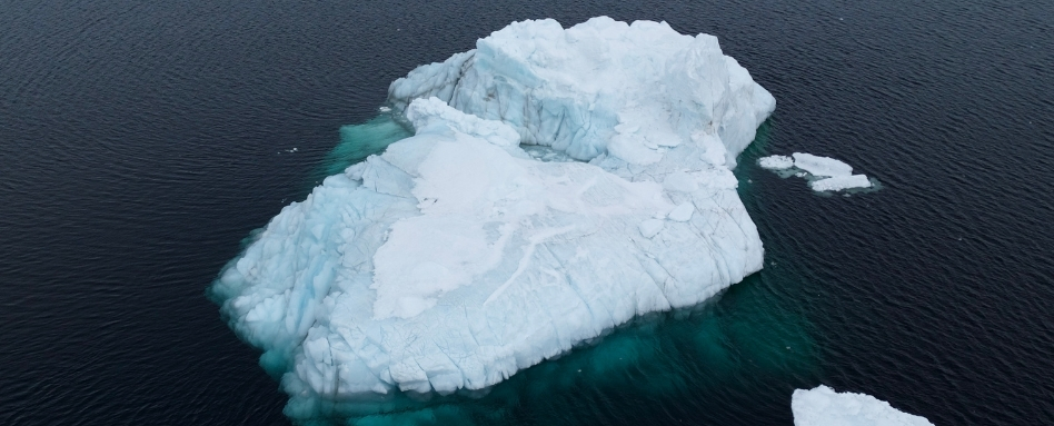 A large iceberg floats in dark blue sea, with turquoise water around its base as seen from above.