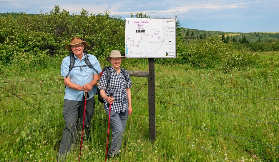 06Support image_SK_Cypress Hills Interprovincial Park_Arlene Robin sign_credit Robin and Arlene Karpan | Trans Canada Trail Two people smiling at the camera, holding hiking poles and standing next to a Trans Canada Trail sign