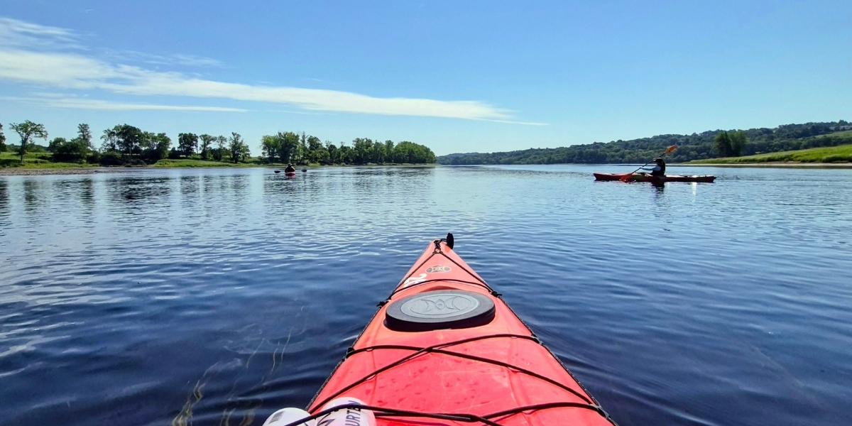00Feature image_Blue Trails NB_credit Cathy Donaldson | Trans Canada Trail Perspective shot from a red kayak on a calm river, with other kayakers in the distance and green banks under a clear blue sky.