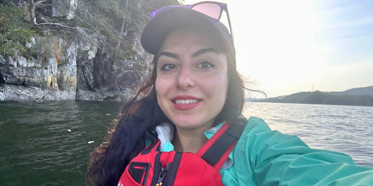 Smiling woman in a red life jacket and teal jacket taking a selfie on a lake, with rocky cliffs and trees in the background.