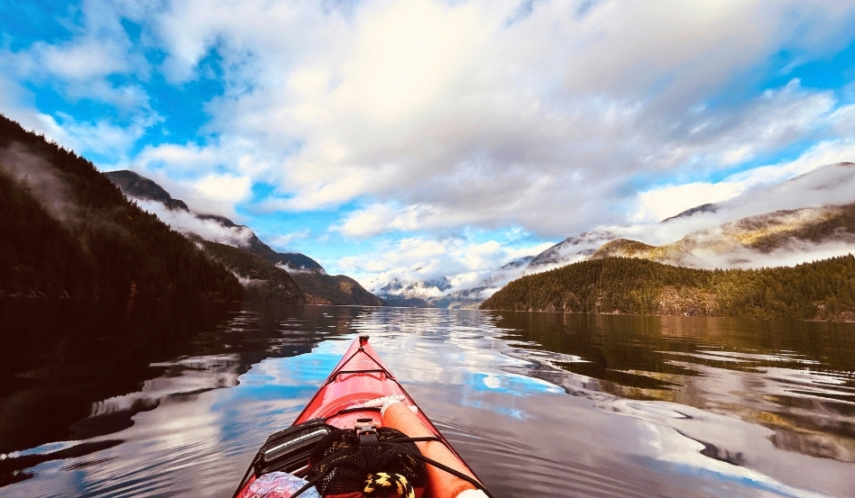Kayaker's red kayak glides across a calm lake surrounded by forested mountains and fluffy clouds in a bright blue sky.