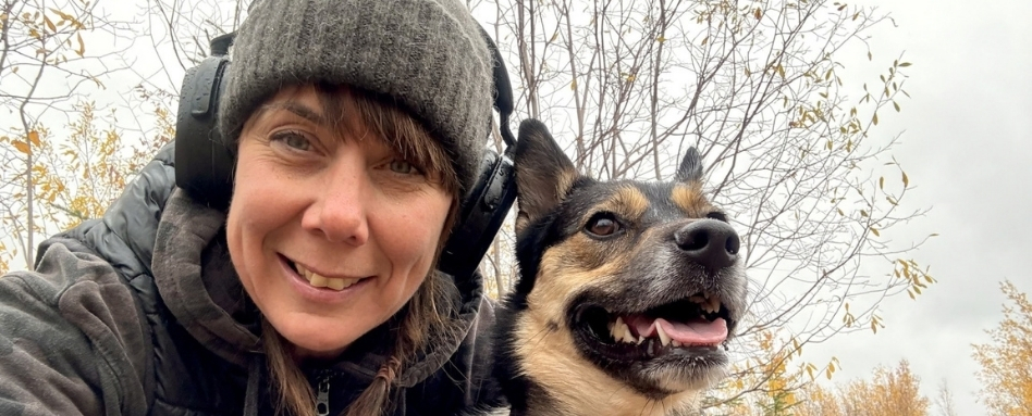 A woman looking at the camera. She is wearing a toque and headphones. Next to her is a dog, who is looking into the distance.