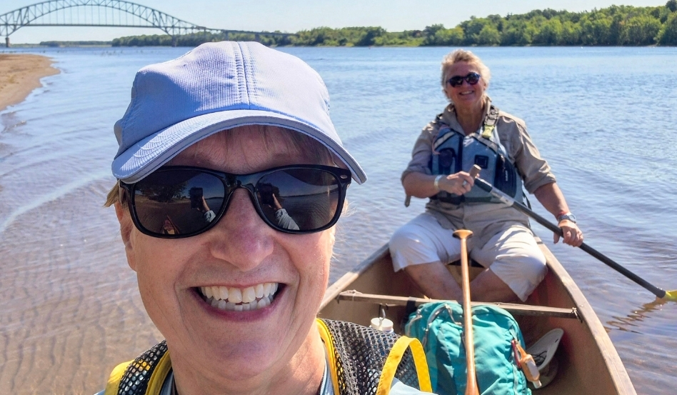 04Support image_Blue Trails NB_credit Cathy Donaldson_Moon and Cathy | Trans Canada Trail Two women paddling a canoe on a sunny river, one in the foreground taking a selfie while the other paddles behind; bridge in the distance.