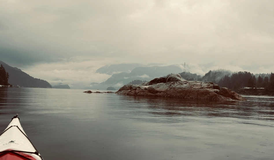 Perspective from a kayak gliding on a calm lake toward a rocky island and misty mountains in the distance, under a cloudy sky, foreground shows part of a kayak.