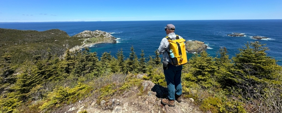 04Support image_NL_web story_East Coast Trail Biscan Cove_credit Taryn Eyton | Trans Canada Trail A person in hiking clothes and a yellow backpack standing on a rocky path, overlooking a forested area and the ocean
