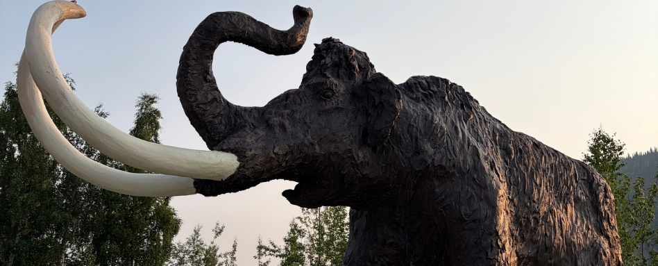 Large mammoth sculpture with curved white tusks outdoors, trees and hills in the background.