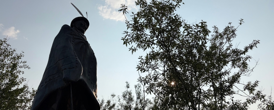 Silhouetted statue of a cloaked warrior holding a sword, set against a pale blue sky and trees behind.
