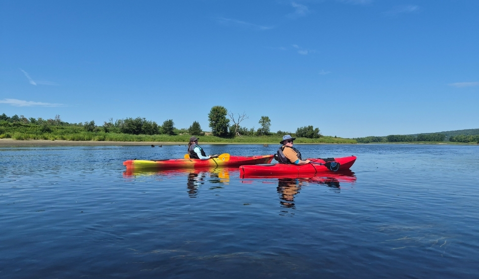 08Support image_Blue Trails NB_credit Cathy Donaldson_Sarah and David | Trans Canada Trail Two people in red and yellow kayaks paddle on a calm river under a clear blue sky and green shoreline.
