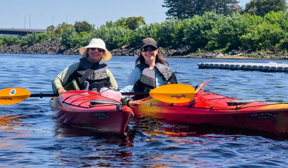 10Support image_Blue Trails NB_credit Cathy Donaldson_duo near end | Trans Canada Trail Two people in red kayaks wearing life jackets and hats, smiling at the camera on calm water with a green shoreline in the background.