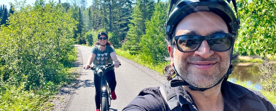 Two adults cycling on a sunny forest trail; man in foreground wearing sunglasses and helmet, woman on bike behind him wearing heart-patterned shirt.