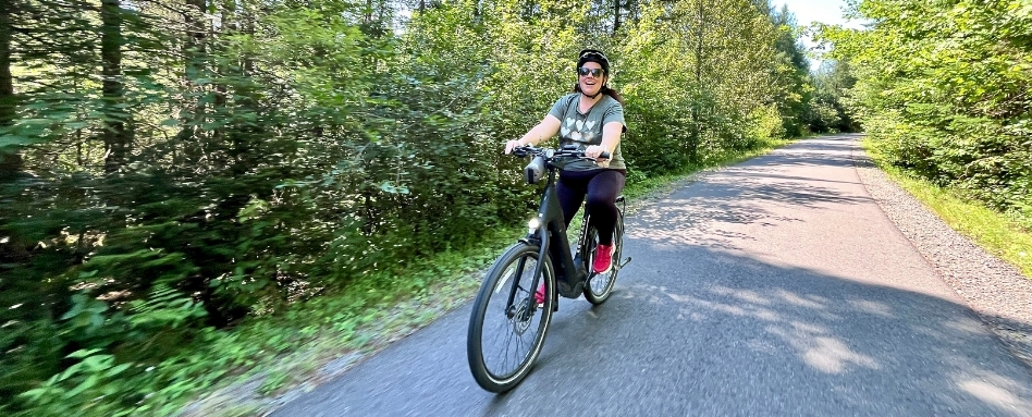 Woman wearing a helmet rides a black bicycle along a tree-lined paved trail in a sunny park area.
