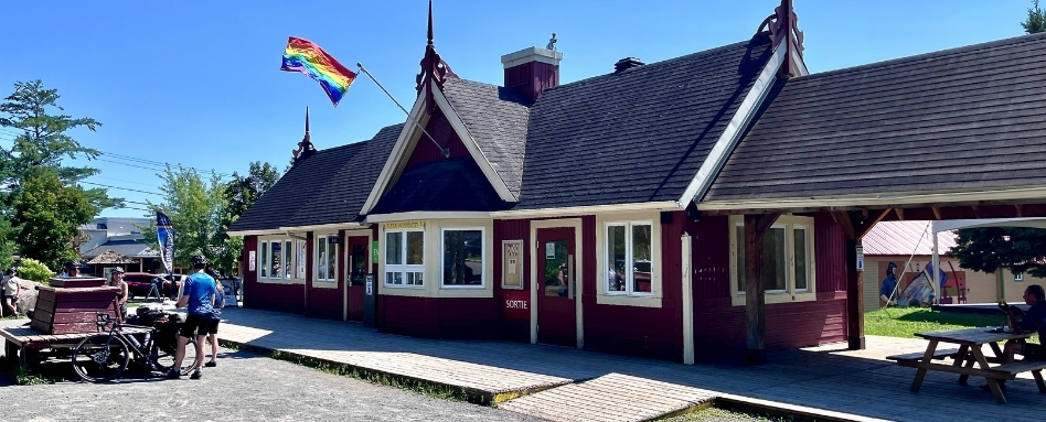 Red wooden station building with a rainbow flag, white-framed windows, and a covered porch; people near bikes and picnic tables outside.