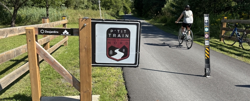 A sign that says P'tit Train du Nord on the left, with a paved trail behind it. A cyclist is riding away from the sign.