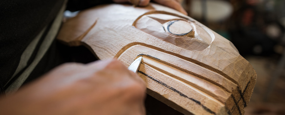 Wood carving by Darren Yelton Squamish Nation artist from North Vancouver British Columbia | Trans Canada Trail Close-up of a woodworker carving a wooden face with a chisel, shaping features on a mask-like sculpture.