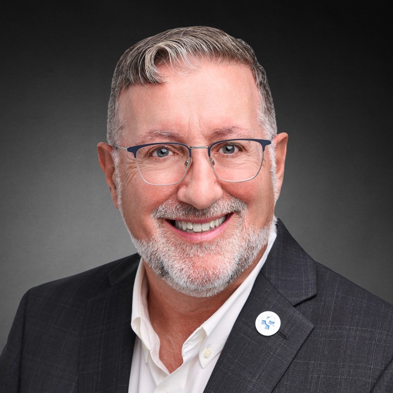Smiling man in a dark blazer and white shirt wearing glasses, with a round lapel pin on his left lapel.