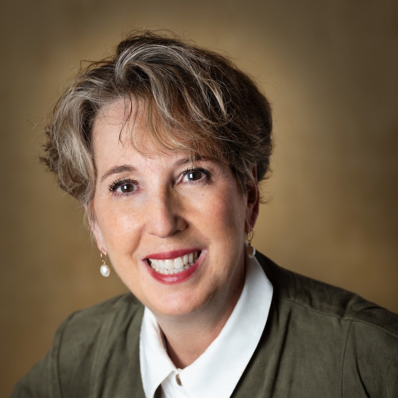 Professional headshot of a smiling woman with short gray hair, wearing a white collar and olive jacket, against a warm brown backdrop.