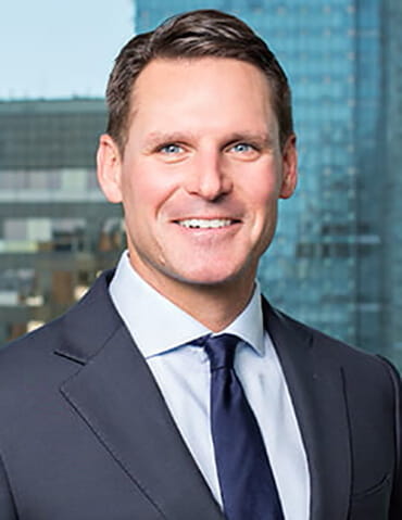 Professional headshot of a smiling man in a dark suit and blue tie, with office buildings in the background.