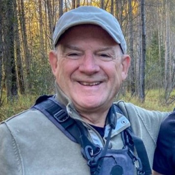 Smiling man wearing a gray cap and outdoor vest, standing in a forested area with trees in the background.