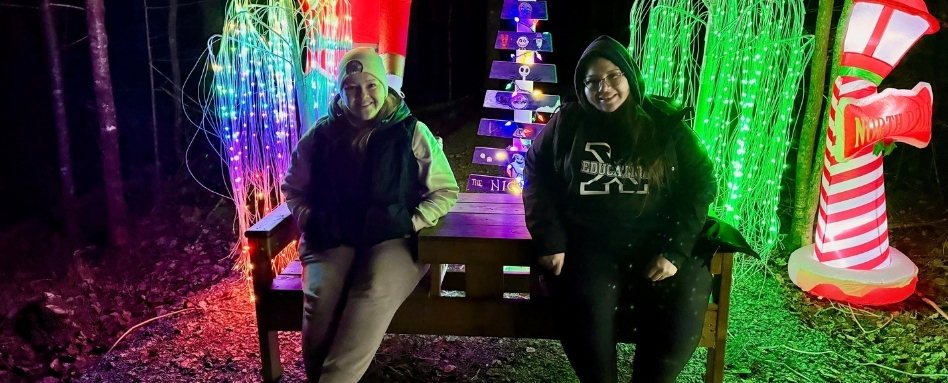 Two people sit on a wooden bench at a nighttime holiday light display, surrounded by multicolored LEDs and a striped inflatable candy cane.