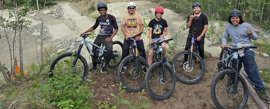 Six riders in helmets posing with mountain bikes on a dirt trail in a wooded area.