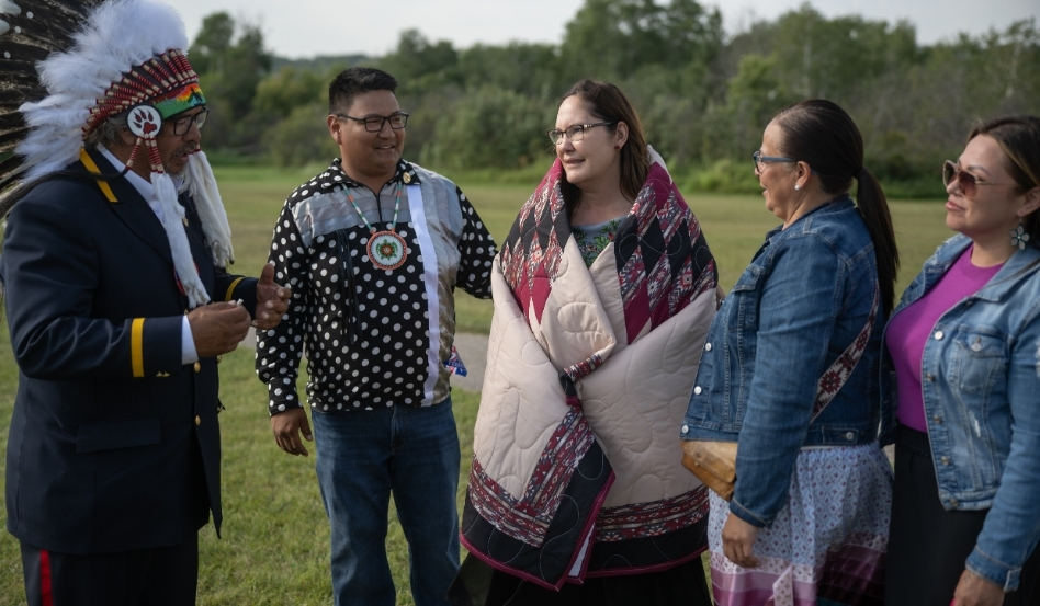 Five people standing together celebrating Treaty 6 Indigenous ceremonies. The woman in the middle has a blanket wrapped around her.