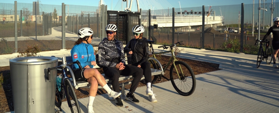 Three cyclists in helmets sit on a bench chatting near their bikes beside a metal trash can and a fence on a sunny paved path.